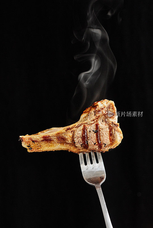 Steaming hot lamb chop, impaled on a fork, held up against a black background